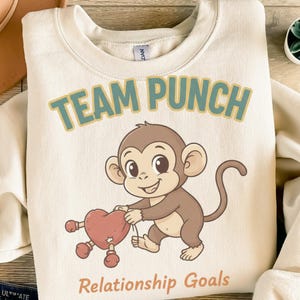 Könnte beinhalten: Cremefarbenes Sweatshirt mit dem Text "TEAM PUNCH" in Türkis und Gelb. Ein Cartoon-Affe hält ein rotes Herz mit Boxhandschuhen. Der Satz "Relationship Goals" ist darunter gedruckt.