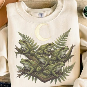 Peut inclure: Sweat-shirt crème avec une illustration détaillée de six grenouilles vertes perchées sur une branche d'arbre, des feuilles de fougère et un croissant de lune. Les grenouilles ont une peau texturée.