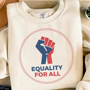Könnte beinhalten: Cremefarbenes Sweatshirt mit einer roten und blauen Faustgrafik in einem roten Kreis, mit den Worten "EQUALITY FOR ALL" in Blau und Rot. Das Sweatshirt ist aus weichem Material und hat einen Rundhalsausschnitt.
