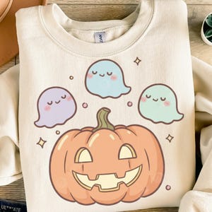 Könnte beinhalten: Cremefarbenes Sweatshirt mit einem lächelnden orangefarbenen Kürbis und drei Cartoon-Geistern in Pastellfarben. Die Geister sind hellblau, lila und grün. Das Design hat ein niedliches, skurriles Halloween-Thema.