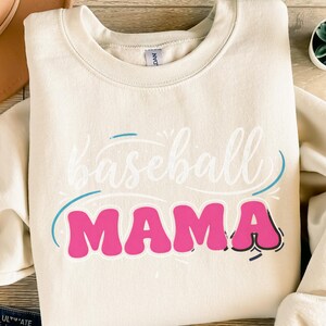 Könnte beinhalten: Cremefarbenes Sweatshirt mit dem Schriftzug "Baseball Mama" in weißer Schrift und pinkfarbenen Buchstaben. Der Text ist blau umrandet. Ein lässiges, bequemes Oberteil für Baseballfans.