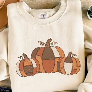 Könnte beinhalten: Cremefarbenes Sweatshirt mit einer Grafik von drei Kürbissen in Braun-, Orange- und Weißtönen. Die Kürbisse haben ein Patchwork-Design und lockige Stiele, ideal für den Herbst.