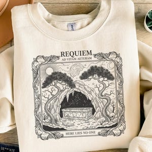 Könnte beinhalten: Cremefarbenes Sweatshirt mit schwarzem Grafikdruck. Das Design zeigt eine Krypta-Szene mit den Worten "REQUIEM AD VITAM AETERAM" und "HERE LIES NO ONE". Die Illustration umfasst Bäume, einen Sarg und einen Schädel.