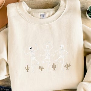 Könnte beinhalten: Cremefarbenes Sweatshirt mit Rundhalsausschnitt. Auf der Vorderseite befindet sich ein gesticktes Design von drei tanzenden Skeletten und Kakteen. Der Text "Fuck Trump" ist zwischen den Skeletten gestickt. Das Sweatshirt ist aus weichem Material.