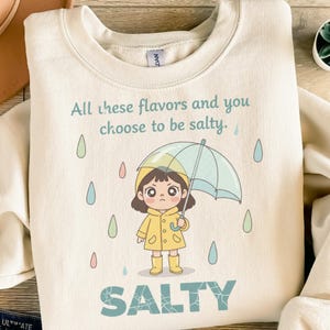 Könnte beinhalten: Cremefarbenes Sweatshirt mit dem Text "All these flavors and you choose to be salty." Ein Cartoon-Mädchen in gelbem Regenmantel und Stiefeln hält einen blauen Regenschirm. Das Wort "SALTY" ist darunter gedruckt.