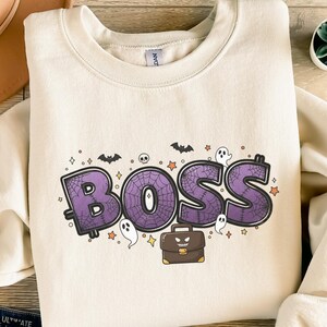 Könnte beinhalten: Cremefarbenes Sweatshirt mit dem Wort "BOSS" in Lila, mit einem Spinnennetz-Design. Das Design umfasst Fledermäuse, Geister, Sterne, einen Totenkopf und eine Aktentasche. Ein Halloween-Themen-Grafik.
