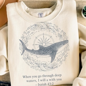 Könnte beinhalten: Cremefarbenes Sweatshirt mit einer Illustration eines blauen Walhais in einem kreisförmigen Design aus Fischen, Wellen und einem Kompass. Der Text "When you go through deep waters, I will a with you - Isaiah 43:2" ist darunter gedruckt.