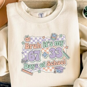 Könnte beinhalten: Cremefarbenes Sweatshirt mit einer farbenfrohen Grafik. Die Grafik lautet "Bruh it's my 67 + 33 Days of School" mit Illustrationen zum Thema Schule. Das Design umfasst ein Schachbrettmuster in Pastellfarben, einen Apfel, einen Rucksack und eine Abschlusskappe.