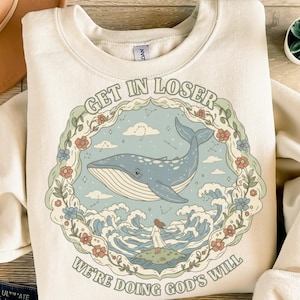 Könnte beinhalten: Cremefarbenes Sweatshirt mit einem kreisförmigen Grafikdesign, das einen Blauwal, Wellen und eine Frau auf einer kleinen Insel zeigt. Der Text "GET IN LOSER" wölbt sich über dem Wal, und "WE'RE DOING GOD'S WILL" steht darunter.