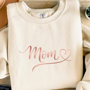 Könnte beinhalten: Cremefarbenes Sweatshirt mit dem Wort "Mom" in roségoldener Schrift, wobei ein Herz den Buchstaben "o" ersetzt. Das Sweatshirt hat einen Rundhalsausschnitt und lange Ärmel. Der Stoff wirkt weich und bequem.
