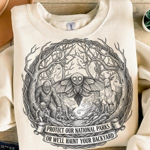 Könnte beinhalten: Cremefarbenes Sweatshirt mit einer Schwarz-Weiß-Grafik von Waldkreaturen, darunter eine Motte, Bigfoot und ein Hirsch, umgeben von Bäumen. Der Text "PROTECT OUR NATIONAL PARKS OR WE'LL HAUNT YOUR BACKYARD" ist auf einem Banner zu sehen.