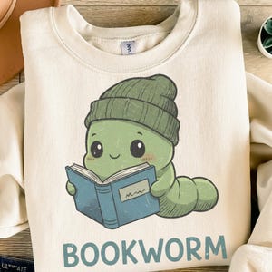 Könnte beinhalten: Cremefarbenes Sweatshirt mit einem Cartoon-Bücherwurm, der eine grüne Mütze trägt und ein blaues Buch liest. Das Wort "BOOKWORM" ist in Großbuchstaben am unteren Rand des Designs aufgedruckt.