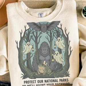Peut inclure: Sweat-shirt crème avec un motif graphique représentant un gorille, une grenouille, des fantômes et une chauve-souris dans un cadre forestier. Le texte "PROTECT OUR NATIONAL PARKS OR WE'LL HAUNT YOUR BACKYARD" est imprimé en bas.