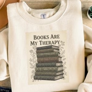 Könnte beinhalten: Cremefarbenes Sweatshirt mit dem Aufdruck "Books Are My Therapy". Die Grafik zeigt einen Stapel Bücher im Vintage-Stil in verschiedenen Farben, umgeben von floralen Illustrationen. Das Sweatshirt hat einen Rundhalsausschnitt und lange Ärmel.