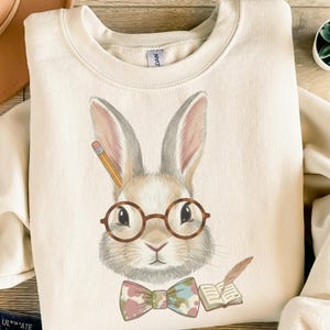 May include: A cream-coloured sweatshirt with a rabbit illustration wearing round glasses and a bow tie. The rabbit holds a pencil, with a book and feather. The design is printed on a soft fabric, suitable for casual wear. The sweatshirt is a comfortable fit.