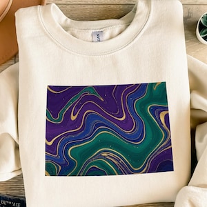Könnte beinhalten: Cremefarbenes Sweatshirt mit rechteckigem Design. Das Design zeigt ein wirbelndes Muster aus Lila, Türkis, Blau und Gold mit Marmoreffekt und goldenen Akzenten. Das Sweatshirt ist aus weichem Material.