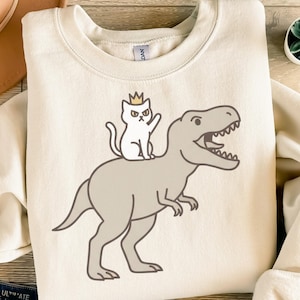 Puede incluir: Sudadera de color crema con una ilustración de dibujos animados. Representa un gato blanco con una corona, montado en un dinosaurio gris. El gato está saludando y el dinosaurio tiene la boca abierta. El diseño está centrado en la parte delantera de la sudadera.