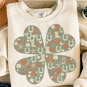 Könnte beinhalten: Cremefarbenes Sweatshirt mit einem vierblättrigen Kleeblatt-Design. Das Kleeblatt besteht aus vier herzförmigen Abschnitten, die jeweils mit Hufeisen-, Cowboy-Stiefel- und Herz-Illustrationen in Orange, Creme und Grün gefüllt sind.