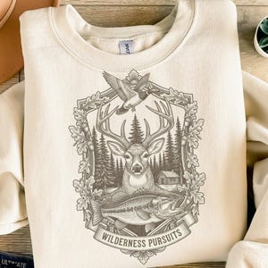Könnte beinhalten: Cremefarbenes Sweatshirt mit detaillierter Grafik eines Hirsches, Fisches und einer Ente in einem dekorativen Rahmen. Das Design umfasst Kiefern und eine kleine Hütte mit dem Schriftzug "Wilderness Pursuits" auf einem Banner.