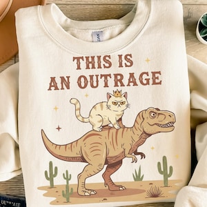 Puede incluir: Sudadera color crema con la frase "THIS IS AN OUTRAGE". El diseño muestra un gato con corona montado en un dinosaurio, con cactus en el fondo. Prenda de estilo casual.