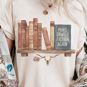 Könnte beinhalten: Cremefarbenes T-Shirt mit einem Bücherregal-Design mit Vintage-Büchern und einem Schädel mit Hörnern. Die Bücher tragen Titel wie "The Good Earth" und "Make Orwell Fiction Again."