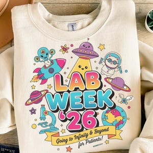May include: Cream-coloured sweatshirt with a colourful graphic design. The design includes a rocket ship, alien, UFO, cat in a spacesuit, and the words "LAB WEEK '26". A banner reads "Going to Infinity & Beyond for Patients!"