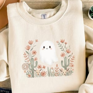 Könnte beinhalten: Cremefarbenes Sweatshirt mit einem skurrilen Geisterdesign. Der Geist ist von einem Blumenarrangement mit Kakteen, Blumen und Schädeln in Rosa-, Grün- und Brauntönen umgeben. Das Sweatshirt hat einen Rundhalsausschnitt und lange Ärmel.