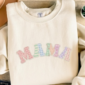 Könnte beinhalten: Cremefarbenes Sweatshirt mit dem Wort "MAMA" in pastellfarbenen Patchwork-Buchstaben. Die Buchstaben sind rosa umrandet und mit Blumen- und Vichy-Mustern versehen. Das Sweatshirt hat einen Rundhalsausschnitt und lange Ärmel.