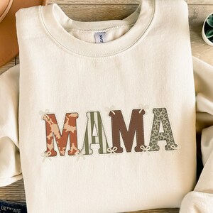 Könnte beinhalten: Cremefarbenes Sweatshirt mit dem Wort "MAMA" in dekorativer Schrift. Die Buchstaben sind mit verschiedenen Mustern versehen, darunter Kuhfellmuster, Streifen und Leopardenmuster. Kleine Schleifen akzentuieren jeden Buchstaben und erzeugen einen gemütlichen, stilvollen Look.