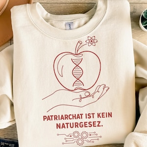 Könnte beinhalten: Cremefarbenes Sweatshirt mit roter Apfelgrafik mit DNA-Strang, gehalten von einer Hand. Der deutsche Text "PATRIARCHAT IST KEIN NATURGESETZ" ist darunter gedruckt, zusammen mit einem Leiterplattendesign.