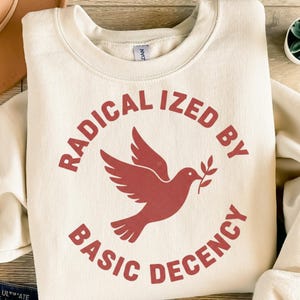 Pode incluir: Camisola creme com decote redondo. A frente apresenta um gráfico vermelho de uma pomba carregando um ramo de oliveira, circundado pelas palavras "RADICALIZED BY BASIC DECENCY" em uma fonte vermelha correspondente. A camisola é feita de um material macio.