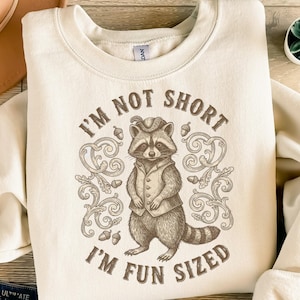 May include: A cream-coloured sweatshirt featuring a raccoon illustration in a waistcoat and hat. The text reads "I'm Not Short, I'm Fun Sized" in brown. The design includes decorative swirls and acorns around the raccoon.