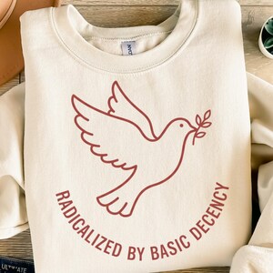 Op de afbeelding: Crèmekleurige sweatshirt met een bordeauxrode omtrek van een duif die een olijftak draagt. De tekst "RADICALIZED BY BASIC DECENCY" is in bordeauxrood rond de duif gedrukt. De sweatshirt heeft een ronde hals.