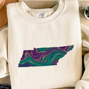 Könnte beinhalten: Cremefarbenes Sweatshirt mit einer Umrisslinie des Bundesstaates Tennessee. Die Umrisslinie ist mit einem wirbelnden Muster aus Teal, Lila und Gold gefüllt. Das Design ist auf dem Rundhals-Sweatshirt zentriert.