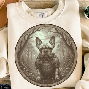 Könnte beinhalten: Cremefarbenes Sweatshirt mit einer detaillierten Illustration einer Französischen Bulldogge in Rüstung, eingebettet in einen kreisförmigen Rahmen, der eine Waldszene darstellt. Das Kunstwerk ist in Braun- und Grautönen gehalten, mit einem Vollmond im Hintergrund.
