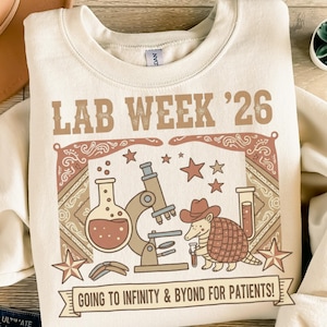 Könnte beinhalten: Cremefarbenes Sweatshirt mit einem Grafikdesign. Das Design zeigt den Text "LAB WEEK '26", ein Mikroskop, Sterne und ein Gürteltier mit Cowboyhut. Das Banner lautet "GOING TO INFINITY & BYOND FOR PATIENTS!"
