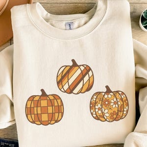 Könnte beinhalten: Cremefarbenes Sweatshirt mit drei Kürbisdesigns in Orange- und Brauntönen. Die Kürbisse haben verschiedene Muster: Karomuster, diagonale Streifen und Blumenmuster. Ein gemütliches Top zum Thema Herbst.