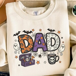 Könnte beinhalten: Cremefarbenes Sweatshirt mit einem Halloween-Motiv "DAD". Die Buchstaben sind mit Fledermäusen, Sternen, Spinnweben und einer Werkzeugkiste verziert. Eine Kaffeetasse mit Totenkopf-Motiv und Geister sind ebenfalls abgebildet.