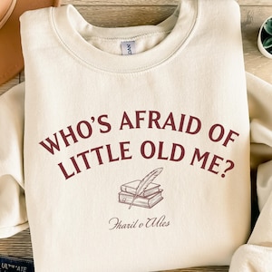 Könnte beinhalten: Cremefarbenes Sweatshirt mit dem burgunderfarbenen Text "WHO'S AFRAID OF LITTLE OLD ME?". Darunter eine Illustration von Büchern und einer Feder. Die Worte "Tharil o Ales" sind unter der Illustration gedruckt.