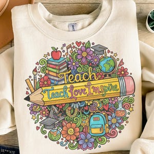 Könnte beinhalten: Cremefarbenes Sweatshirt mit einem farbenfrohen Grafikdesign. Das Design zeigt einen großen gelben Bleistift mit den Worten "Teach Love Inspire" und andere schulbezogene Elemente wie Bücher, Blumen und einen Globus. Das Sweatshirt hat einen Rundhalsausschnitt.