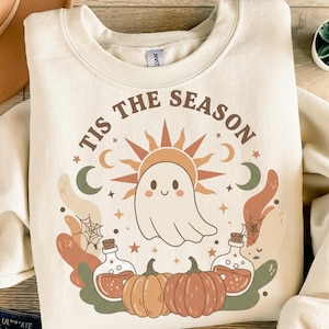Könnte beinhalten: Cremefarbenes Sweatshirt mit einem Halloween-Motiv. Das Design zeigt einen lächelnden Geist, eine Sonne, Kürbisse, Zaubertränke und den Text "TIS THE SEASON" in kreisförmiger Anordnung. Die Gesamtästhetik ist herbstlich.