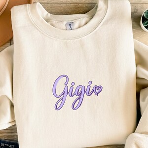 Peut inclure: Sweat-shirt crème avec le nom "Gigis" en écriture cursive lavande. Le mot a un point en forme de cœur au-dessus de la lettre "i". Le sweat-shirt est fait d'une matière douce.