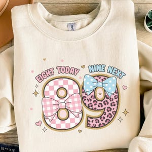 Könnte beinhalten: Cremefarbenes Sweatshirt mit dem Text "EIGHT TODAY NINE NEXT" und der Zahl "89". Die "8" ist rosa kariert mit einer rosa Schleife, und die "9" ist Leopardenmuster mit einer blauen Schleife. Kleine Herzen und Sterne sind verstreut.