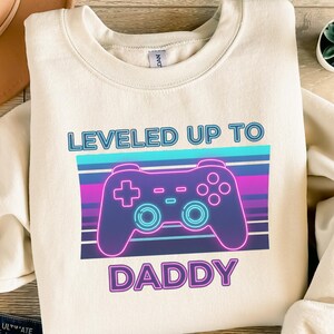 Könnte beinhalten: Cremefarbenes Sweatshirt mit einem Grafikdesign eines Videospiel-Controllers in Lila, Blau und Rosa. Der Text "LEVELED UP TO DADDY" ist oberhalb und unterhalb des Controller-Designs gedruckt.
