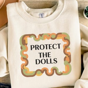 Könnte beinhalten: Cremefarbenes Sweatshirt mit dem Text "PROTECT THE DOLLS" in Schwarz, umrahmt von einem Retro-Rahmen in Braun-, Grün- und Orangetönen. Das Sweatshirt hat einen Rundhalsausschnitt und lange Ärmel.