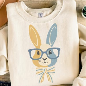 Könnte beinhalten: Cremefarbenes Sweatshirt mit einem skurrilen Hasen-Design. Der Hase hat gelbe und blaue Ohren, trägt eine blaue Brille und eine passende Fliege. Die Grafik ist auf der Vorderseite des langärmeligen Rundhalsausschnitts zentriert.