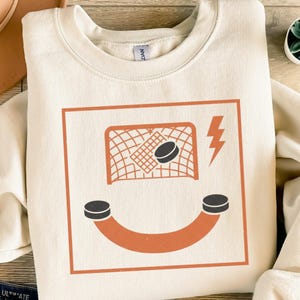 Könnte beinhalten: Cremefarbenes Sweatshirt mit einem Hockey-Motiv. Das Design umfasst ein Hockeyspielnetz mit einem Puck, zwei Pucks, die ein Lächeln bilden, und einen Blitz. Die Grafik ist in Orange- und Schwarztönen gehalten.
