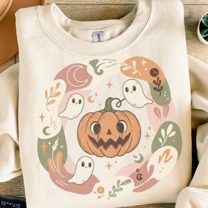 Könnte beinhalten: Cremefarbenes Sweatshirt mit einem Halloween-Motiv. Das Design zeigt einen lächelnden orangefarbenen Kürbis, freundliche weiße Geister, Halbmonde und florale Elemente in gedämpften Farben. Das Sweatshirt hat einen Rundhalsausschnitt.
