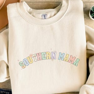 Könnte beinhalten: Cremefarbenes Sweatshirt mit dem Text "SOUTHERN MAMA" in pastellfarbenen Karobuchstaben. Das Sweatshirt hat einen Rundhalsausschnitt und lange Ärmel. Der Text ist über der Brust gebogen.