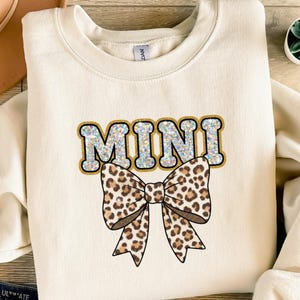 Può includere: Felpa color crema con la scritta "MINI" in un carattere glitterato, azzurro chiaro, bordato d'oro. Sotto il testo c'è un grande fiocco con un disegno a stampa leopardata marrone e beige. La felpa ha uno scollo tondo.